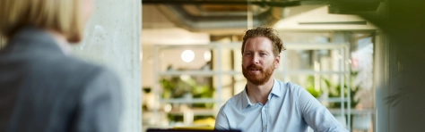 Ginger man with beard sitting down in smart blue shirt talking to business woman in grey suit and blonde hair. Public and social sector manager having meeting on bright office environment.