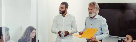 due colleghi parlano in piedi davanti ad un tavolo di altri colleghi e colleghe