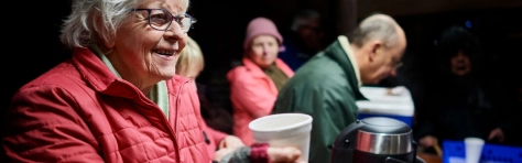 Mature female in warm clothing, serving hot drink at local voluntary soup kitchen