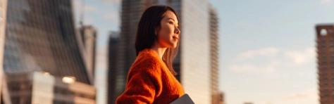 Thoughtful young Asian carrying laptop, looking at view in financial district at sunset.