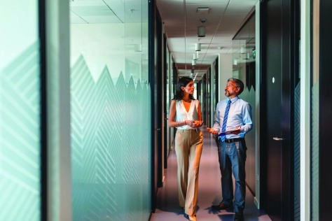 A man and a woman are walking and talking together down a long, modern office corridor. The woman, on the left, has dark hair and is wearing a sleeveless white blouse with beige wide-leg trousers, holding a laptop or folder under her arm. The man, on the right, has grey hair and a beard and is dressed in a light blue dress shirt with a blue patterned tie and grey trousers, gesturing with his hand mid-conversation while holding documents. The corridor features frosted glass partitions with a decorative geometric/mountain pattern, dark door frames, and overhead recessed lighting, giving the scene a sleek, professional corporate atmosphere.