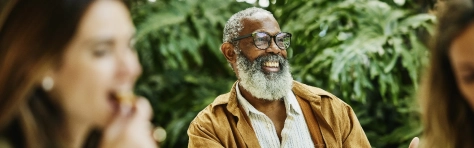 Senior and mature man sitting down smiling in a social environment. Man smiling with large grey beard and smart glasses and woman in the foreground eating food.