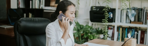 woman using phone with her customer while working on computer laptop at the greenery office in the morning
