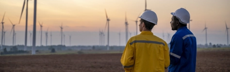 Two environmental engineers standing in a field with hard hats on and PPE. Background is on a wind turbine farm.