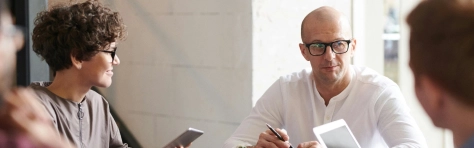 Office workers sitting down around table discussing business. Man with glasses talking to peers in an office setting. HR team, payroll services, employees.