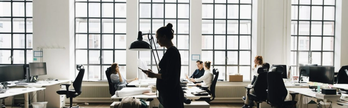 donna in primo piano con telefono in mano che cammina in un open space con altri colleghi che lavorano dietro di lei